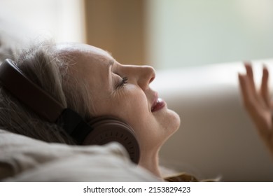 Happy serene senior grey haired lady in headphones listening to relaxing chill out tunes, classical music concert for relaxation, mindfulness, meditation, resting on sofa, smiling with closed eyes