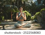 Happy serene young Hispanic woman meditating doing yoga breathing exercises feeling mental mind balance sitting in lotus pose hands in namaste in peaceful green nature park outdoors.