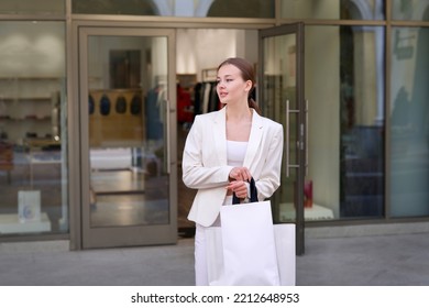 Happy Serious Elegant Stylish Girl, Young Teen Teenager Beautiful Woman In White Is Smiling In Mall Outside Shop, Store Or Boutique, Shopping, Carrying Shopping Paper Bags In Hand 