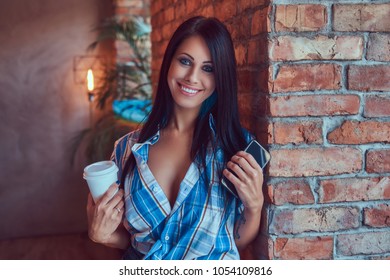 A Happy Sexy Brunette In A Flannel Shirt And Jeans Holds A Cup Of Coffee And The Phone Posing Against A Brick Wall. 