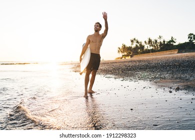 Happy Shirtless Male Surfer In Shorts With Sunblock On Face Standing On Seashore With Surfing Board Under Arm And Waving Hand On Sunny Evening At Tropical Resort