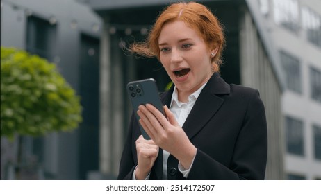 Happy shocked excited Caucasian ginger woman female girl businesswoman formal suit celebrate victory achievement holding mobile phone good news win message rejoice business success outside city street - Powered by Shutterstock