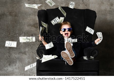 Happy shouting self-confident rich kid boy millionaire sitting with legs crossed in luxury armchair and throwing money dollars cash. Overspend.