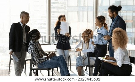 Happy shy young african american employee holding paper documents in hands, presenting marketing research results to interested multiracial diverse colleagues at brainstorming meeting in office.