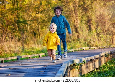 Happy Siblings Having Fun Running On Wood Path Walk In Sunny Autumn Forest, Playing Catch Up