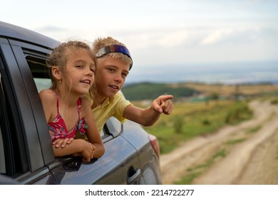 Happy Siblings Looking Out From Window At Car And Enjoying Road Trip. Boy And Girl Admire View From Car Window And Boy In Sunglasses Points His Hand Into The Distance