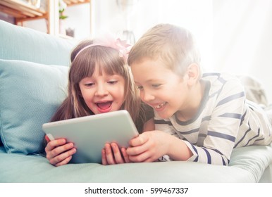 Happy siblings lying on sofa at home and playing with pc tablet together