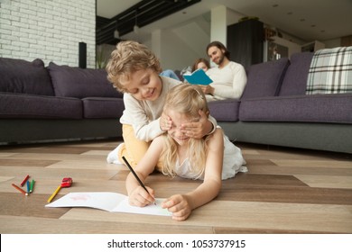 Happy siblings playing peek a boo or guess who drawing on warm wooden floor while parents reading book on sofa, kid brother covering sisters eyes smiling having fun together at home in living room