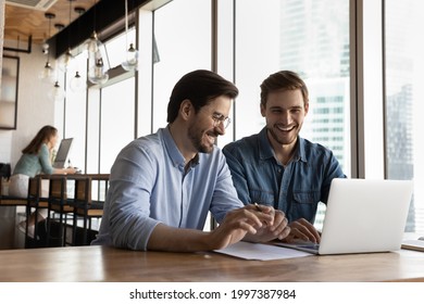 Happy sincere friendly young male colleagues laughing, holding pleasant conversation or watching funny video, enjoying break pause timer working on online project on computer in modern office room. - Shutterstock ID 1997387984