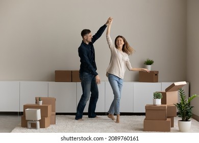 Happy Sincere Young Family Couple Dancing To Music In Living Room Between Carton Parcels. Joyful Millennial Spouses Homeowners Celebrating Moving Day In New Apartment, Real Estate Service Concept.