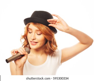 Happy Singing Girl. Beauty Woman Wearing White T-shirt And Black Hat With Microphone Over White Background. Hipster Style.