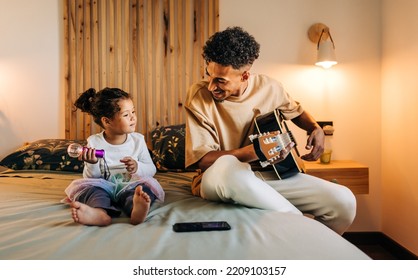 Happy single dad smiling at his daughter while playing a guitar at home. Cheerful dad and daughter having a good time while sitting on a bed together. Father and daughter spending some quality time. - Powered by Shutterstock