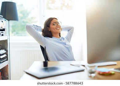 Happy single woman in long sleeve shirt and eyeglasses with arms folded behind head while seated at desk