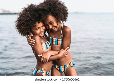 Happy sisters embracing inside sea water during summer time - Afro kids having fun playing on the beach - Family love and travel vacation lifestyle concept 