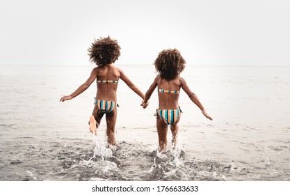 Happy Sisters Running Inside Water During Summer Time - Afro Kids Having Fun Playing On The Beach - Family Love And Travel Vacation Lifestyle Concept 