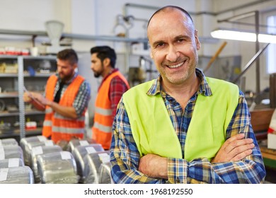 Happy skilled worker stands in front of the warehouse of a metal factory with a team in the background