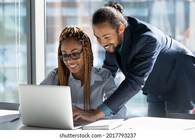 Happy skilled young male african american boss leader manager helping smiling female biracial colleague with computer software applications, working together on online project in modern workplace.