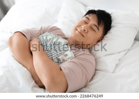 Happy sleeping middle aged chinese man hugging bunch of cash dollars, lying on bed, asian male keeps money safe, top view. Savings, financial literacy, economy concept