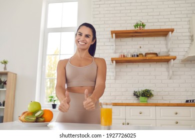 Happy Slender Female Athlete Raises Thumbs Up Showing That Fresh Fruit Juice And Fruit Is Super. Woman Stands In The Kitchen After A Morning Workout And Encourages Everyone To Drink Vitamin Juice.