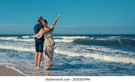 A happy slender romantic middle-aged couple enjoying a relaxing walk on a sandy beach on a sunny day, against the background of large sea waves. - Powered by Shutterstock