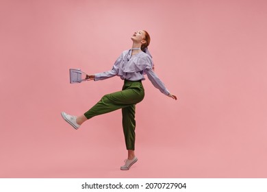 Happy slim young model waving her arms and legs with her head tilted back in isolated studio room. Energetic fair-skinned lady with red hair in lilac blouse, green pants. Good mood, fashion trends
