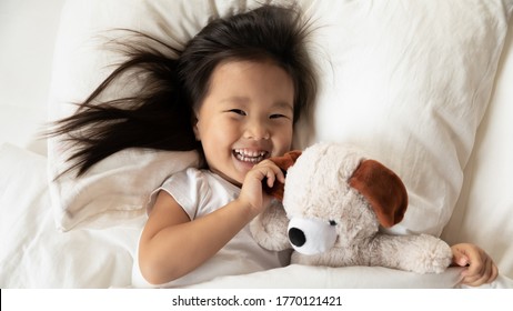 Happy Small Adorable Vietnamese Kid Girl Lying On Pillow Under Duvet, Looking At Camera, Feeling Energetic After Waking Up In Morning, Playing With Toy Staying In Bed, Above Top Head Shot Close Up.