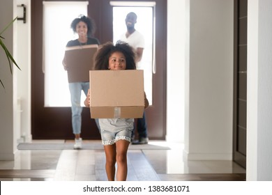 Happy small African American girl holding cardboard box run into new house, young black family moving into bought apartment, excited daughter carry personal belongings to home. Relocation concept