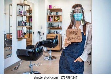 Happy Small Business Owner At A Hairdressing Studio Hanging An Open Sign During COVID-19. Portrait Of Elegant Hair Salon Employee In Apron With Medical Mask, Gloves, Hair Comb And Scissors.