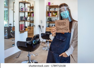 Happy small business owner at a hairdressing studio hanging an open sign during COVID-19. Portrait of elegant hair salon employee in apron with medical mask, gloves, hair comb and scissors. - Powered by Shutterstock