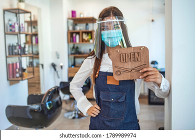 Happy Small Business Owner At A Hairdressing Studio Hanging An Open Sign During COVID-19. Portrait Of Elegant Hair Salon Employee In Apron With Medical Mask, Gloves, Hair Comb And Scissors.