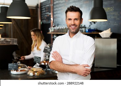 Happy small business owner standing at front of bar with employee in background preparing coffee