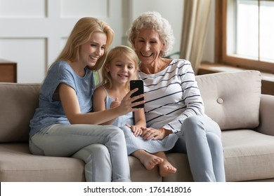 Happy small girl with young mother and aged grandmother at home, multi-generational family sit on sofa in living room use cell, enjoy distant talk via video call, making self-portrait photo for memory