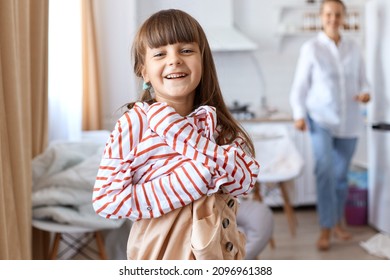 Happy Small Kid With Dark Hair Wearing Large Striped Shirt And Beige Short Pretending To Be A Grown Up Female, Looking At Camera With Happy Expression, Posing With Mother And Kitchen Set On Background