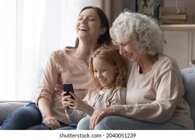 Happy small kid girl holding cellphone in hands, laughing at funny photo video content with joyful young mother and older senior retired granny, relaxing together at home, tech addiction concept. - Powered by Shutterstock