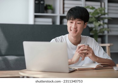 A happy and smart young Asian male college student in casual clothes is doing his homework at a coffee table in his living room. - Shutterstock ID 2311340753