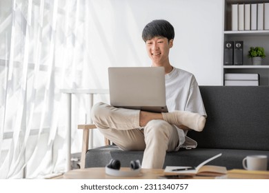 A happy and smart young Asian male college student in casual clothes is doing his homework at a coffee table in his living room. - Shutterstock ID 2311340757