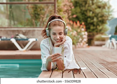 Happy Smartphone Woman Relaxing Near Swimming Pool Listening With Earbuds To Streaming Music. Beautiful Girl Using Her Mobile Phone 