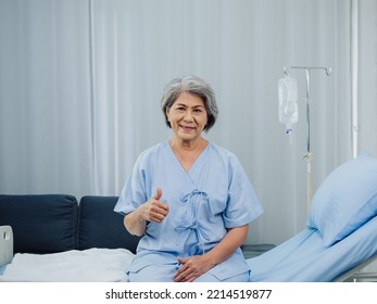 Happy smile Asian elderly old female patient with grey hair in light blue dress thumb up while sitting on bed near the medical IV pole stand with saline drip in white room background in the hospital. - Powered by Shutterstock