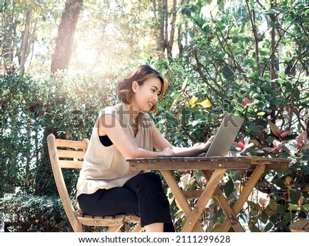 Happy smile Asian woman short hair in beige sleeveless shirt sit and working with laptop computer on wood table, relaxing at the outdoor, surrounded with green plants in the garden. Work everywhere.