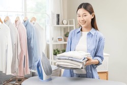 Happy Smile Asian Young Woman Holding Stack, Pile Folded Hygiene Laundry Clothes At Ironing Board After Use Electric Iron Steam At Home. Housekeeping Lifestyle, Household Of Chores, Housework Concept.