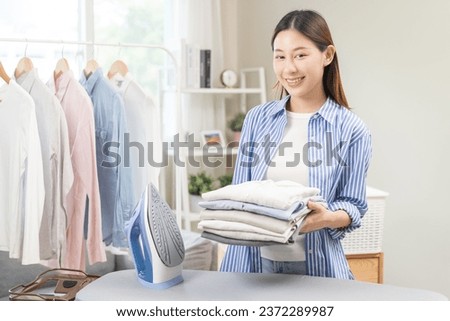 Happy smile asian young woman holding stack, pile folded hygiene laundry clothes at ironing board after use electric iron steam at home. Housekeeping lifestyle, household of chores, housework concept.