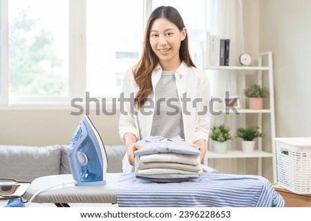 Happy smile asian young woman holding stack, pile folded hygiene laundry clothes at ironing board after use electric iron steam at home. Housekeeping lifestyle, household of chores, housework concept.