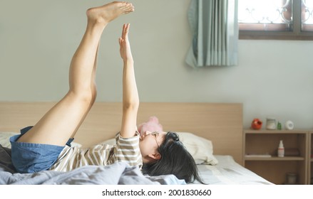 Happy Smile Portrait Young Adult Asian Woman Exercise Leg Up In Air Rest On Bed Wear Eyeglasses. People Stay At Home Lifestyle Concept.