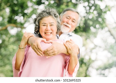 Happy Smile Senior Asian Couple Enjoying Quality Time At The Park And Making Heart Sign. Focus On Hand