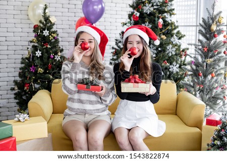 Happy smile of two young Asian women dressing up for theme Christmas with holding gifts box and red ball covering one's nose at home in celebrating Christmas festival together in happy holidays.