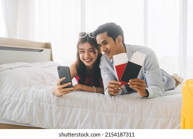 Happy Smile Young Adult Southeast Asian Couple Using Mobile Phone For Getting Ready For Holidays Travel Trip. Living At Home Laying On Bed Together. Curtain On Background With Day Light