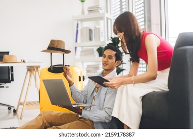 Happy Smile Young Adult Southeast Asian Couple Using Laptop For Getting Ready For Holidays Travel Trip. Living At Home Sitting On Sofa With Hat And Luggage. Window Day Light Background.