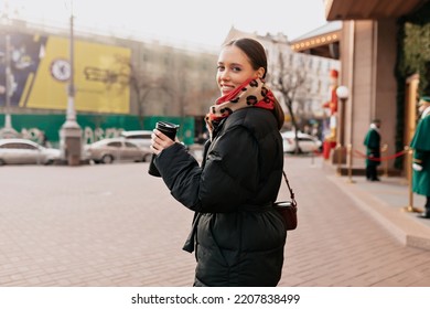 Happy Smiling Adorable Girl With Dark Hair Wearing Black Jacket And Bright Scarf Turn Around At Camera With Happy Smile. She Drinks Coffee And Enjoys Sunny Day