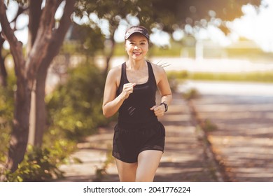 Happy Smiling Adult Asian Woman Jogging Outdoor In The City Park In Sunshine Beautiful Summer Day.  Happy Relaxed Mature Woman Jogging To Live An Active And Healthy Lifestyle