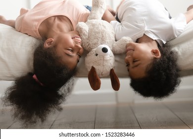 Happy smiling african american children boy and girl with soft toy laying upside down on bed, joking, having fun, looking at each other, fooling at home, enjoying free playtime together. - Powered by Shutterstock